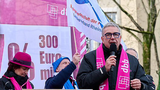 Redner mit Handmikrofon auf einer Bühne unter freiem Himmel vor gewerkschaftlichem Kampagnenlogo