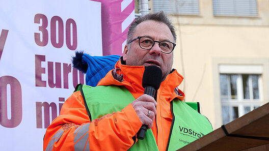 Mann mit Handmikrofon an einem Rednerpult unter freiem Himmel