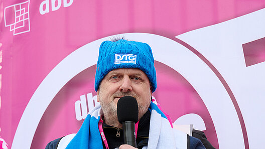 Mann mit Handmikrofon vor einem Kampagnenlogo.