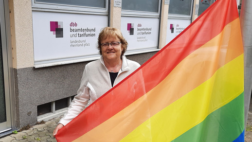 Frau hält Zipfel einer gehissten Regenborgenfahne und steht vor Bürogebäudefassade mit dbb Logo