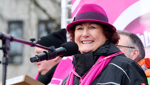 Frau mit magentafarbenem Hut an einem Mikrofon unter freiem Himmel